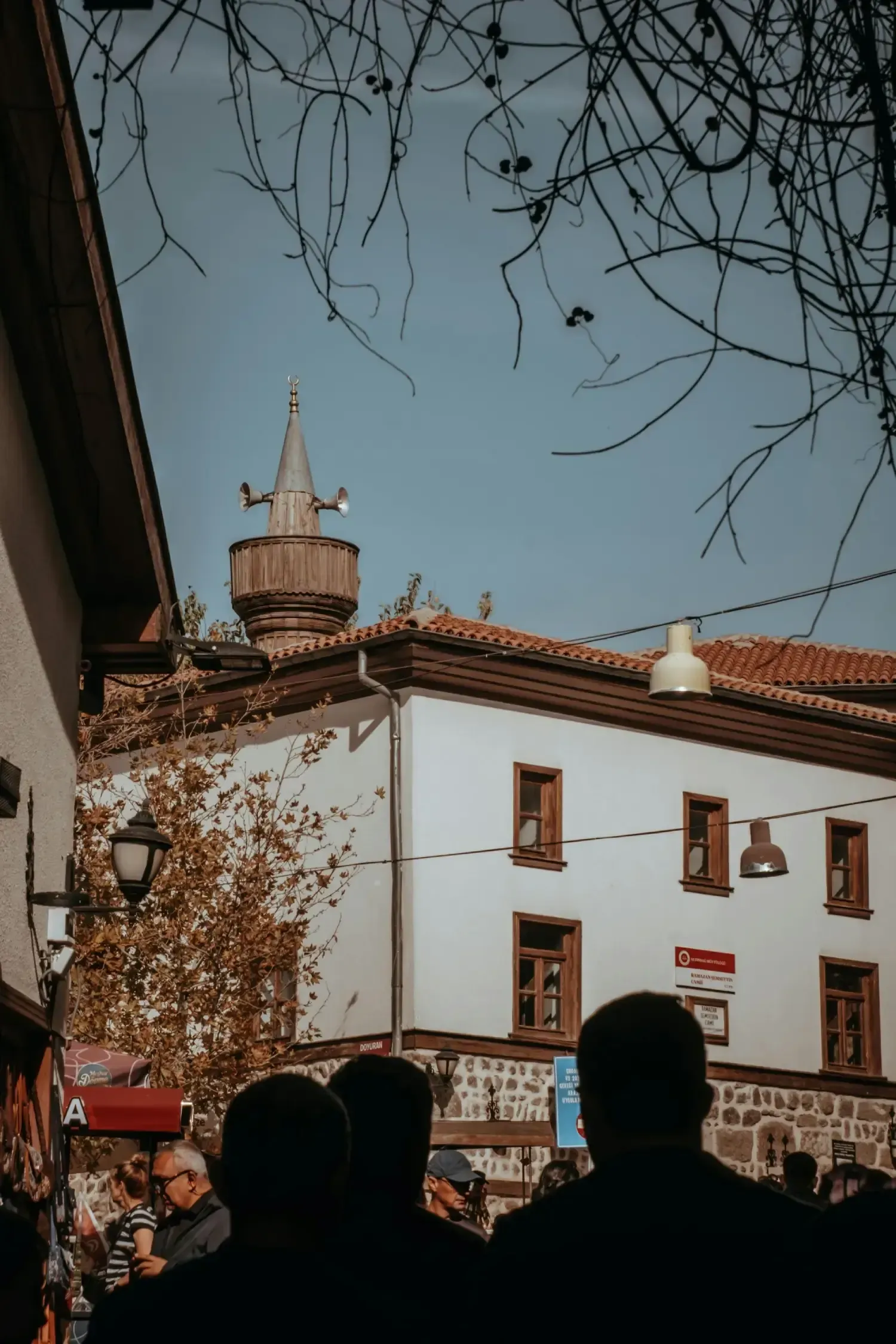 Edificios con minarete, ramas de árboles desnudos y figuras en silueta bajo un cielo azul.