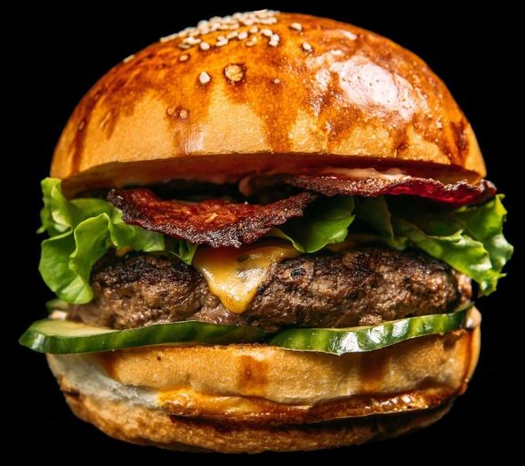 Hamburguesa con semillas de sésamo, lechuga, tocino, queso fundido y pepinillos sobre un fondo negro.