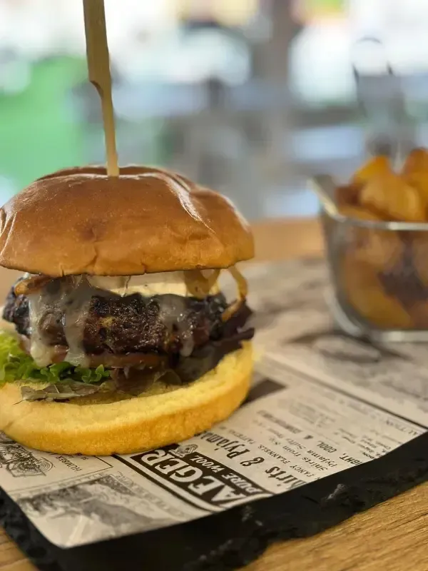 Hamburguesa en una bandeja con patatas fritas al fondo, servida en papel tipo periódico en un restaurante.