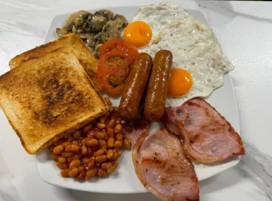 Plato de desayuno con tostadas, judías al horno, huevos, salchicha, beicon, champiñones, tomate y tomates cherry.