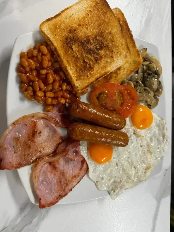 Plato de desayuno con tostadas, frijoles, jamón, salchichas, huevos fritos, champiñones y rodajas de tomate.