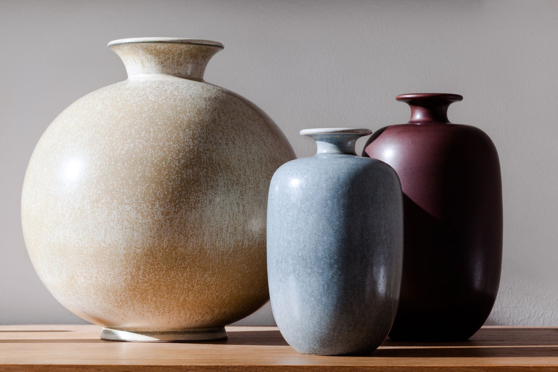 Trois vases en céramique, beige, bleu et bordeaux, sont posés sur une surface en bois devant un fond neutre.