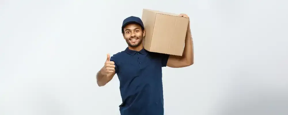 A delivery man is holding a cardboard box on his shoulder and giving a thumbs up.