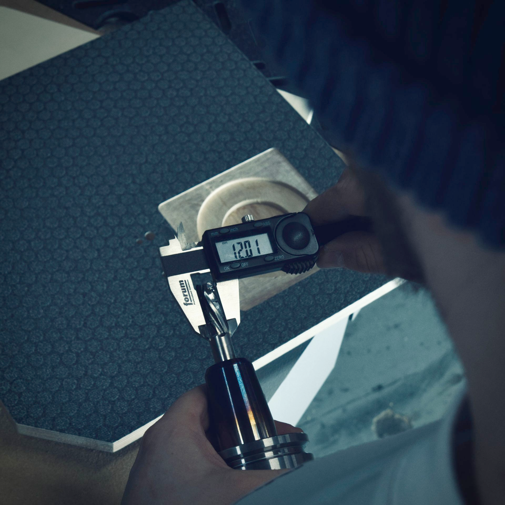 Eine Person misst mit einem digitalen Messschieber ein Objekt auf einer Arbeitsfläche. Das Display zeigt 12,01 an.