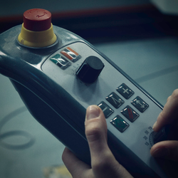 Eine Hand bedient ein graues industrielles Bedienfeld mit Knöpfen, einem Joystick und einem roten Not-Aus-Knopf.