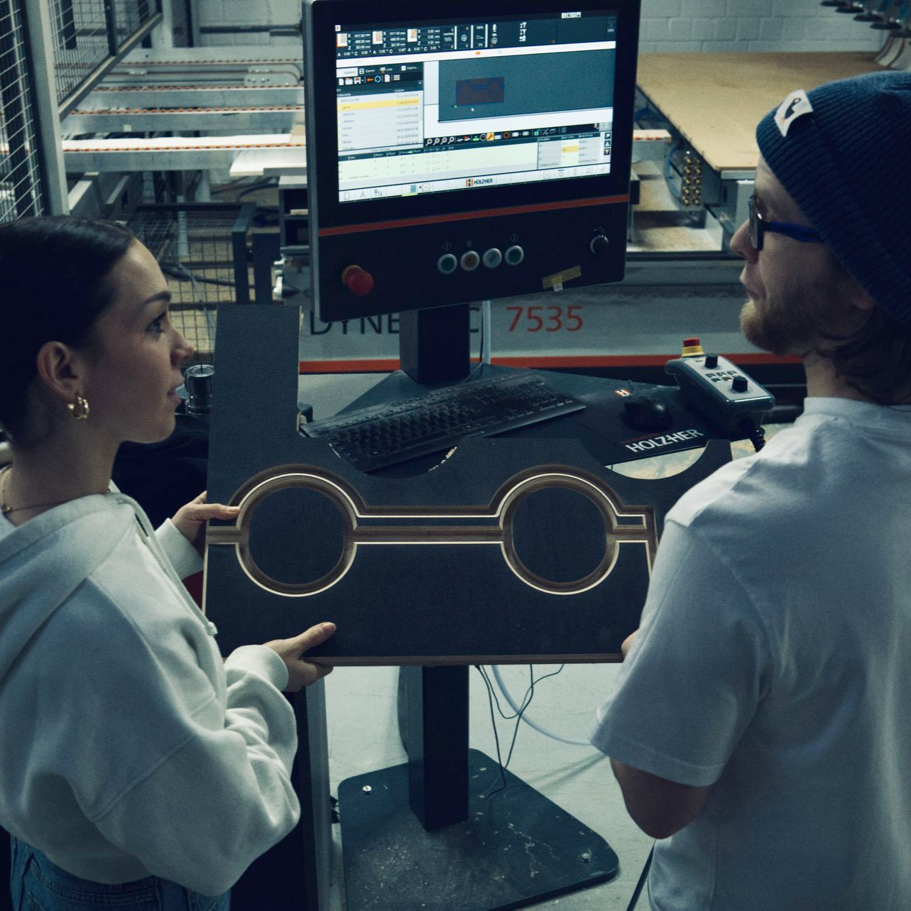 Zwei Personen betrachten ein dunkles, geometrisches Holzstück in der Nähe einer CNC-Maschine in einer Werkstatt.