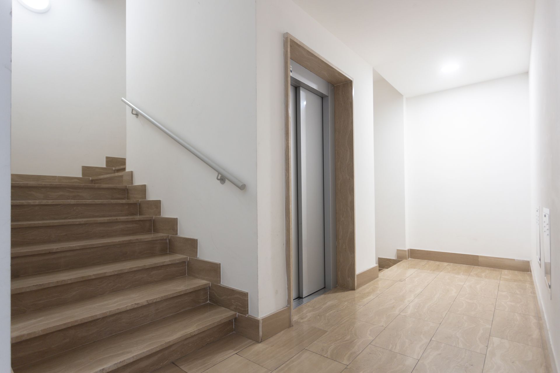 Escalier et ascenseur dans un couloir avec sol carrelé beige et murs blancs ; rampe d'escalier.