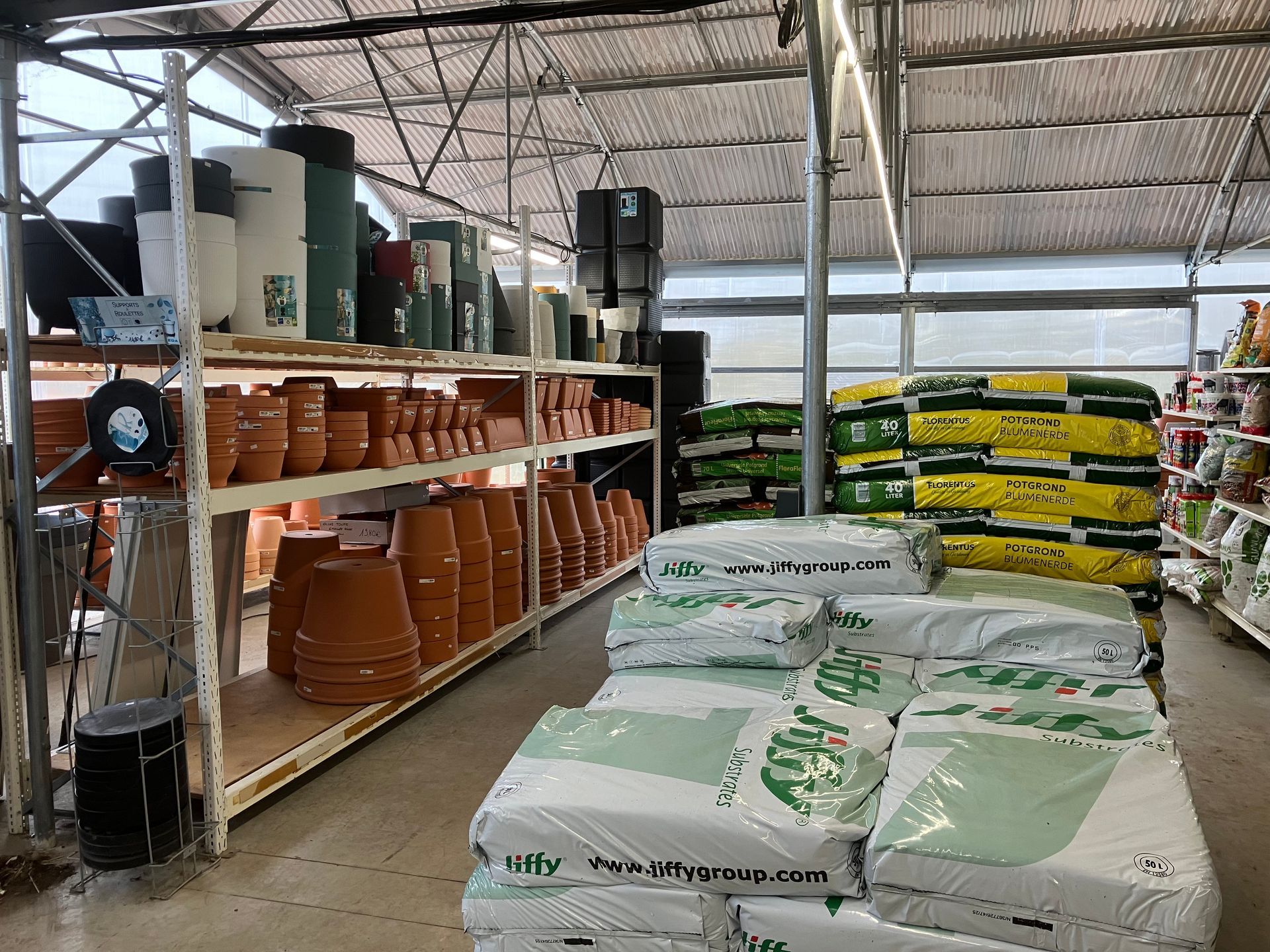 Une jardinerie avec des pots en terre cuite empilés sur des étagères et des sacs de terreau posés au sol sous une serre.