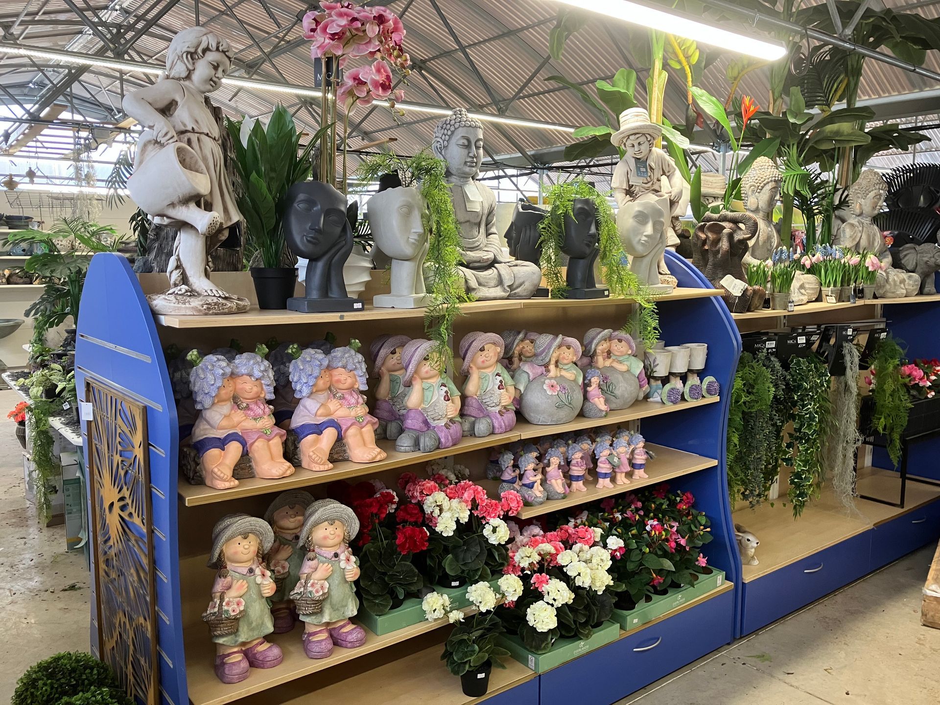 Dans une jardinerie, des étagères présentent des figurines décoratives pour le jardin, des plantes en pot et des fleurs artificielles.