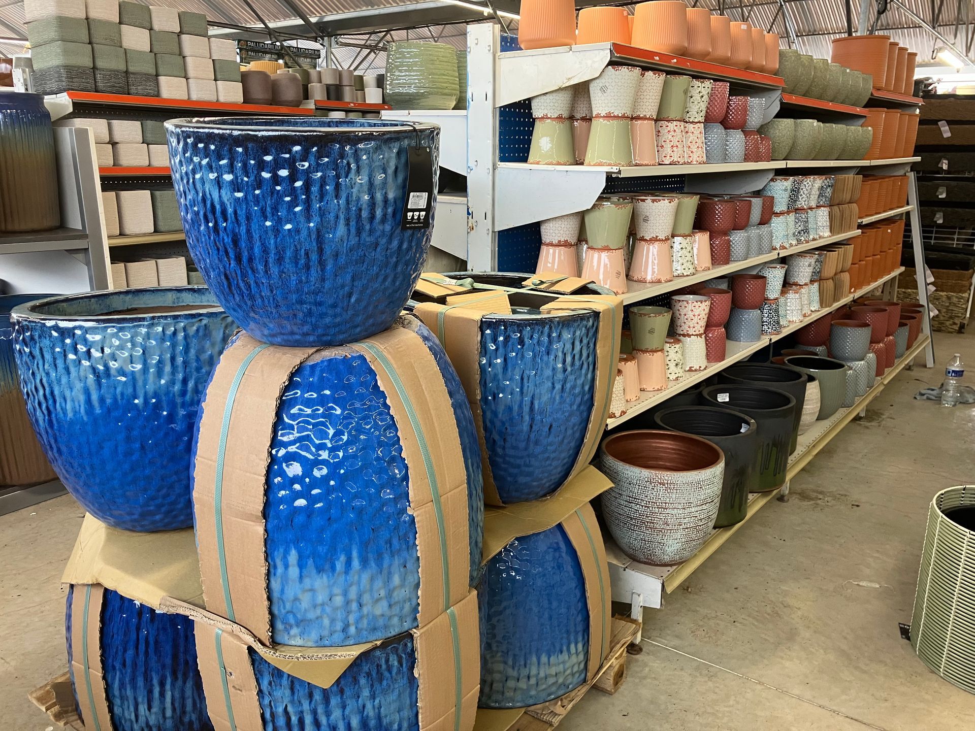 Des pots de fleurs en céramique bleu vif sont empilés dans une allée d'un magasin de jardinage remplie de poteries diverses sur des étagères métalliques.