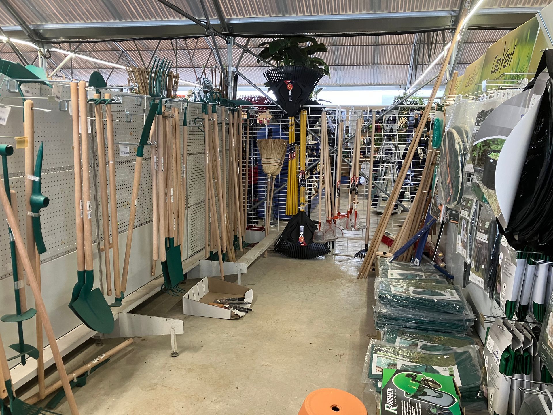 Une allée d'une jardinerie remplie d'outils en métal vert à long manche, de pelles et de râteaux, présentés sur des étagères murales.
