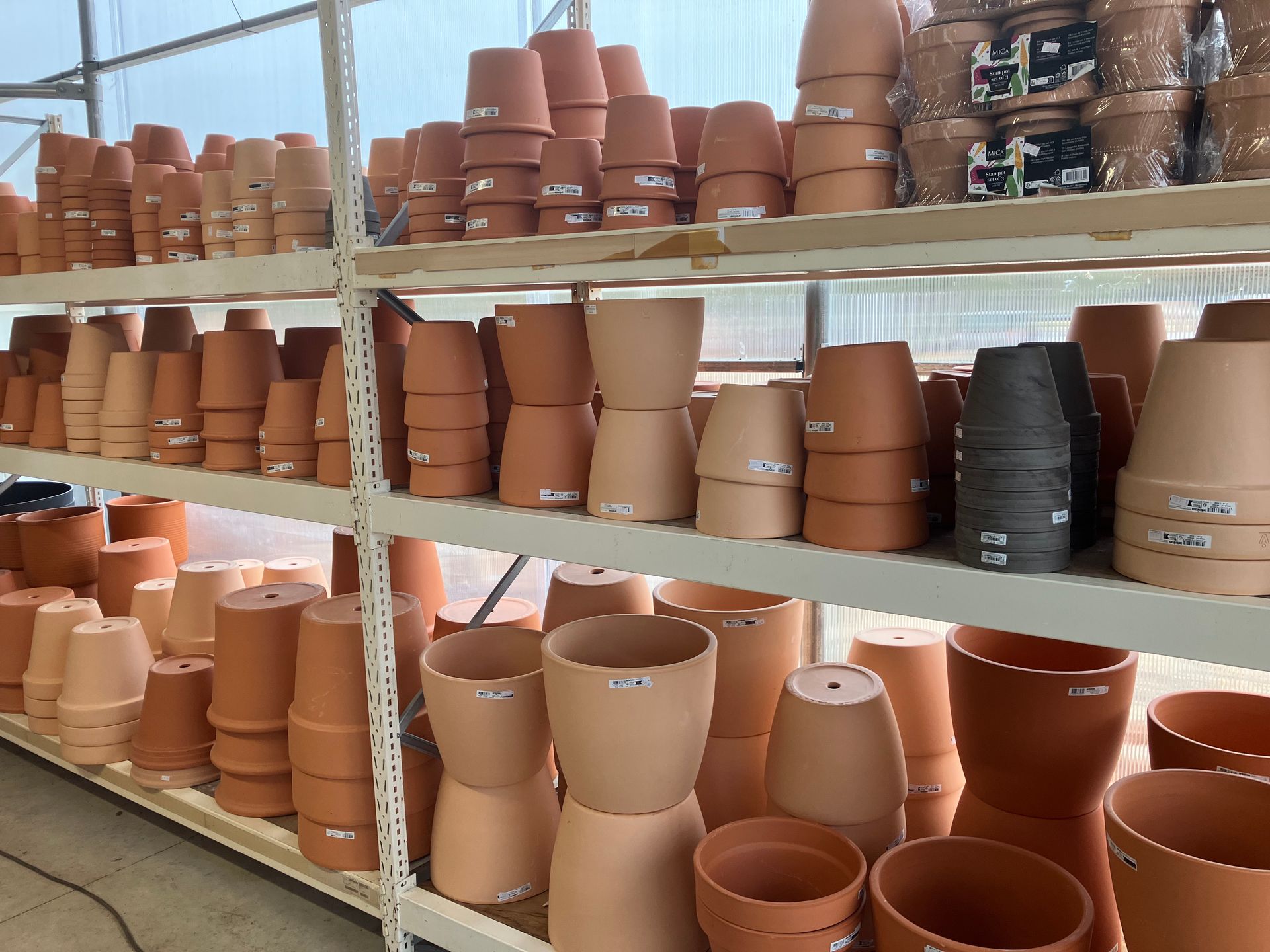 Des étagères dans une serre remplies de rangées de pots de fleurs en terre cuite et en argile grise de différentes tailles, empilés les uns sur les autres.