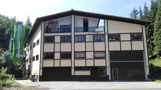 Edificio beige de dos plantas, con detalles oscuros y balcón, situado junto a árboles altos en una zona rural.