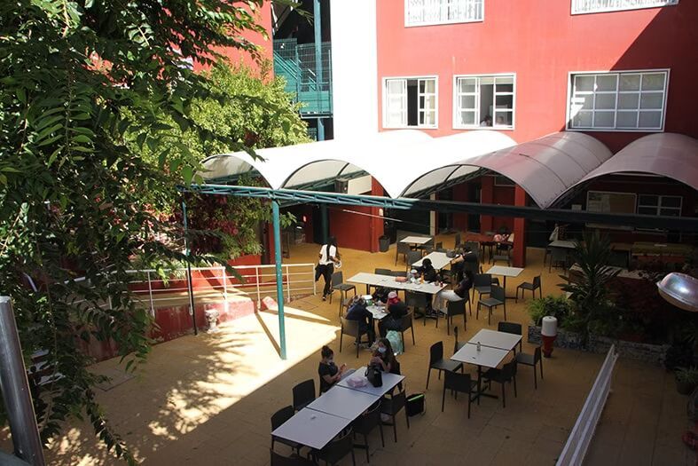Área de refeições ao ar livre no pátio, com mesas e pessoas sob toldos brancos ao lado de um edifício vermelho.