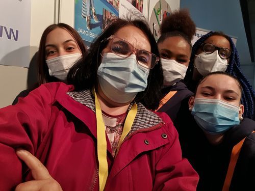 Cinco amigos usando máscaras posam para uma selfie em um ambiente interno, com uma jaqueta vermelha em primeiro plano.