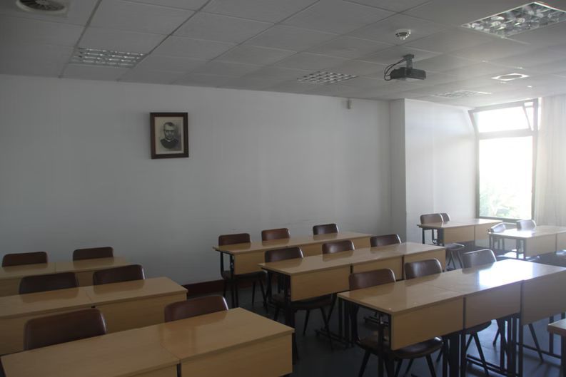 Sala de aula vazia com carteiras de madeira, paredes brancas e luz solar intensa entrando por uma janela.