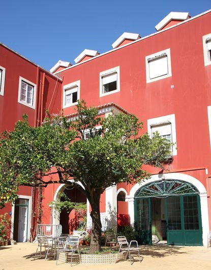 Prédio vermelho com arcos adornados de branco, árvores e mesas de café ao ar livre em um pátio ensolarado.