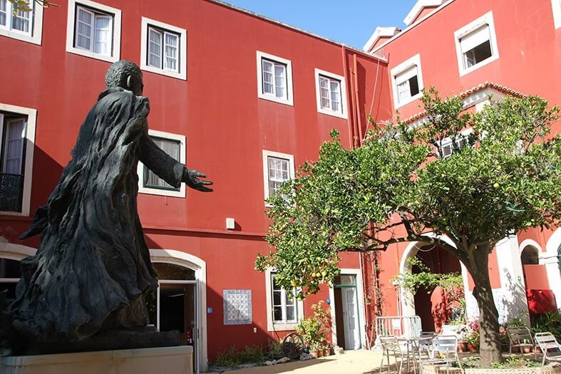 Estátua de bronze em um pátio vermelho com janelas de moldura branca e um arco de árvores frondosas.