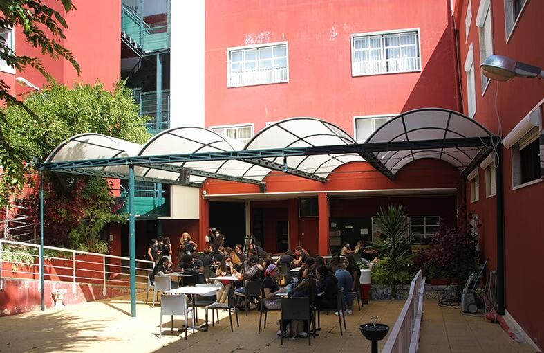 Pessoas sentadas à mesa em um pátio ensolarado entre prédios vermelhos sob uma cobertura de vidro.