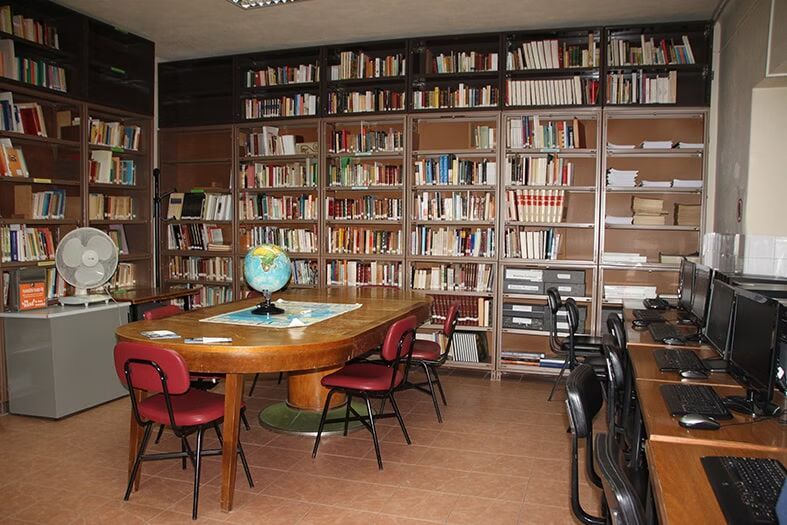 Sala de biblioteca com estantes de livros, mesa redonda, cadeiras e computadores ao longo da parede direita.