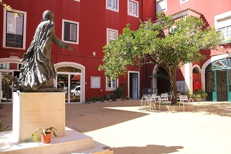 Pátio com edifícios vermelhos, uma estátua de bronze sobre um pedestal e uma árvore que sombreia mesas ao ar livre.