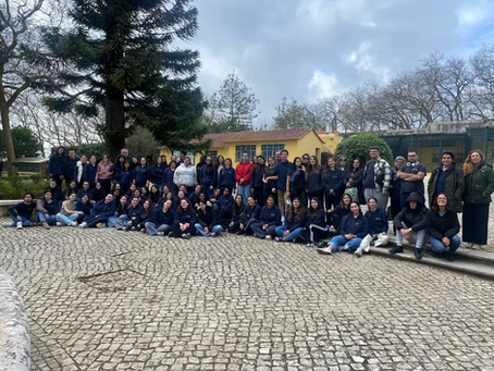 Um grande grupo posando ao ar livre em um piso de paralelepípedos em frente a um prédio amarelo e árvores.