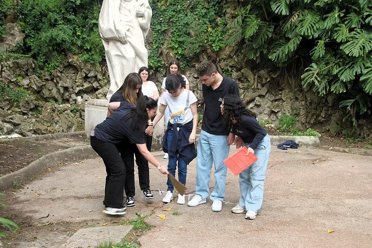 Quatro pessoas ao ar livre perto de uma estátua, apontando para o chão em um jardim verdejante.