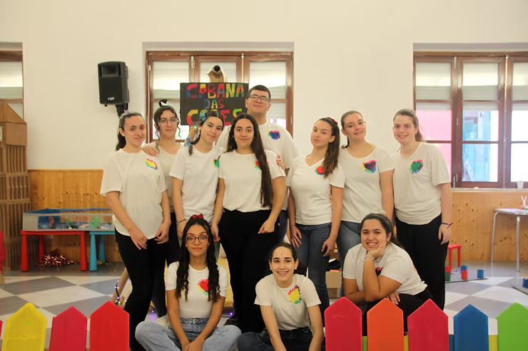 Grupo de pessoas posando em um ambiente interno em frente a blocos coloridos e janelas, vestindo camisas brancas e calças pretas.