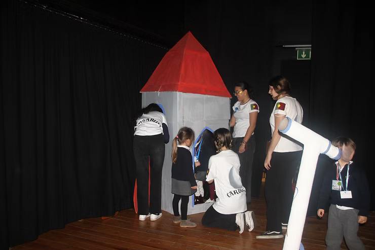 Crianças e adultos em um quarto escuro montando uma casa de papelão com telhado vermelho (modelo cenográfico).