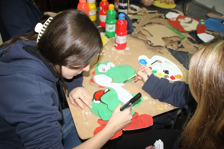 Duas crianças pintando recortes de Natal em uma mesa com frascos de tinta coloridos por perto.