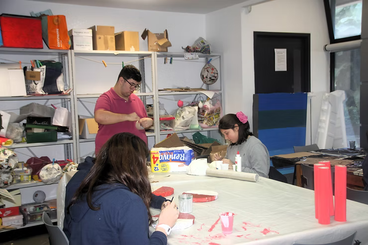 Pessoas sentadas ao redor de uma mesa fazendo artesanato em uma sala desarrumada.