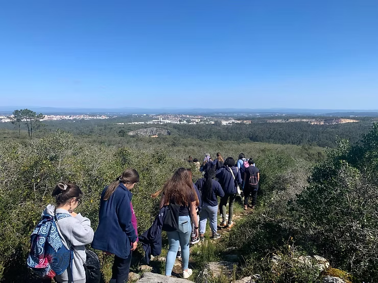 Grupo caminhando por uma trilha rochosa através de colinas verdejantes, com uma cidade ao longe sob um céu azul claro.