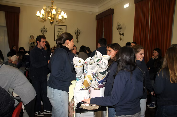 Pessoas reunidas em torno de uma mesa com objetos empilhados em uma sala aconchegante com cortinas e lustres.