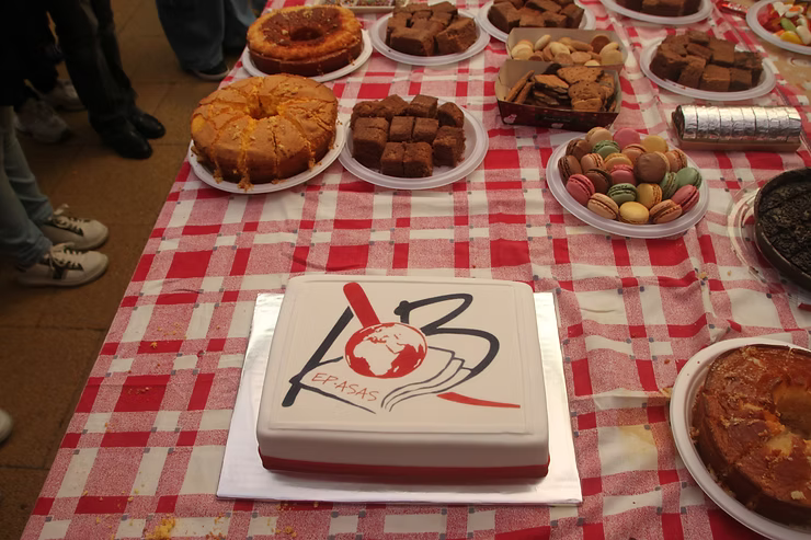 Mesa de sobremesas com toalha xadrez, bolos, biscoitos e um bolo quadrado decorado no centro.