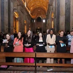 Visitantes em pé atrás de uma barreira de madeira dentro de uma grande catedral, segurando panfletos e olhando para a frente.