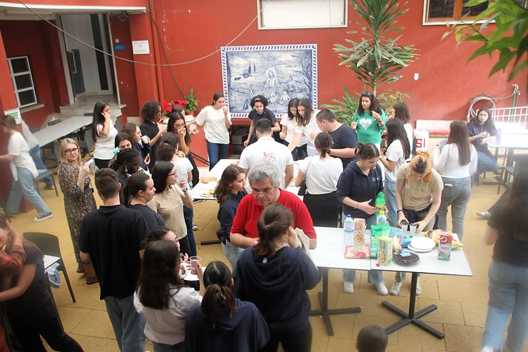 Em um pátio murado de vermelho, pessoas se reuniam em volta de mesas, conversando e servindo comida em um evento informal.