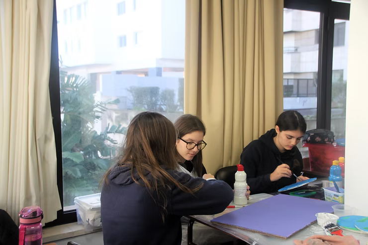 Três pessoas sentadas à mesa perto de uma janela, trabalhando com papéis e cadernos.
