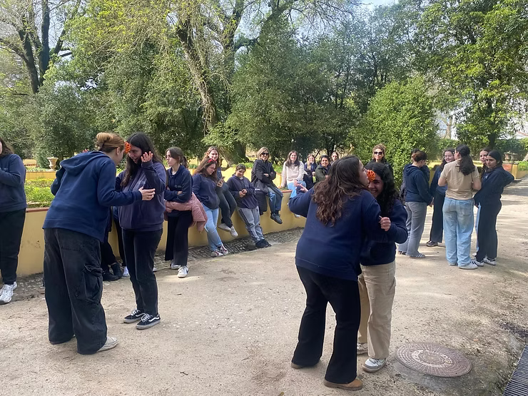 Pessoas em pé em um parque arborizado, algumas conversando e outras frente a frente em pequenos grupos.