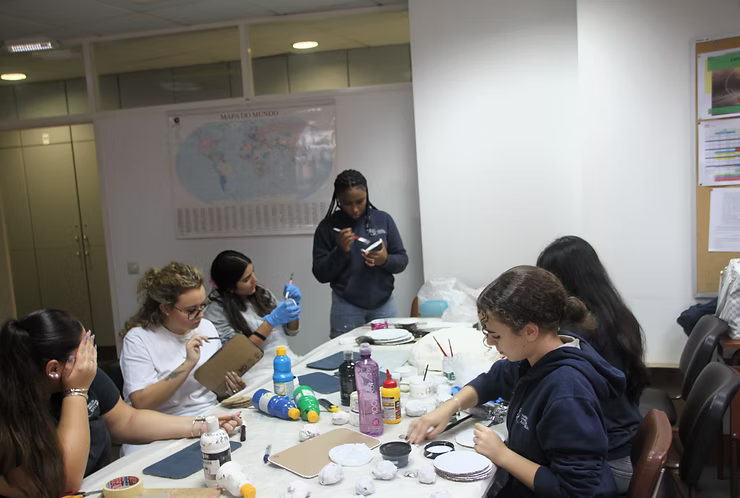 Pessoas sentadas ao redor de uma mesa fazendo trabalhos manuais em uma sala de aula ou sala de reuniões.