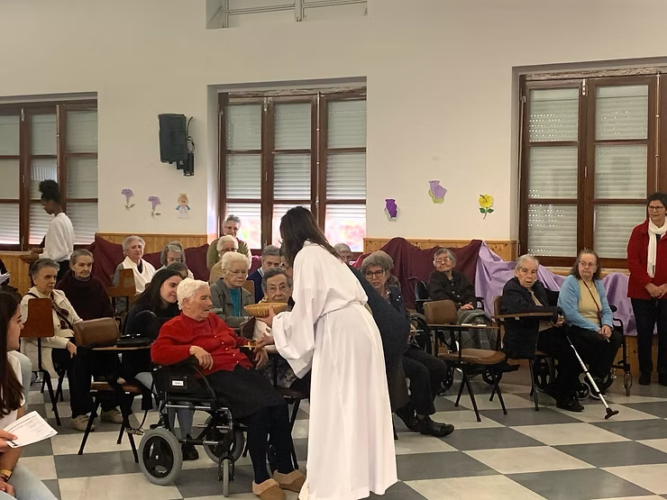 Mulher vestida de branco conversando com moradores sentados em uma sala comunitária com piso quadriculado e janelas.