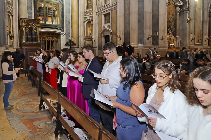 Pessoas em pé numa igreja durante um culto, algumas lendo folhetos, com bancos e colunas ornamentadas por perto.