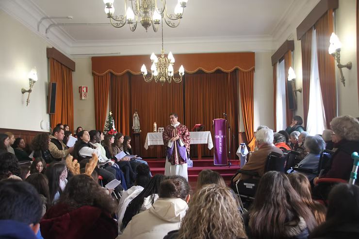 Público sentado em um salão assistindo a um palestrante em um palco com cortinas roxas e lustres.
