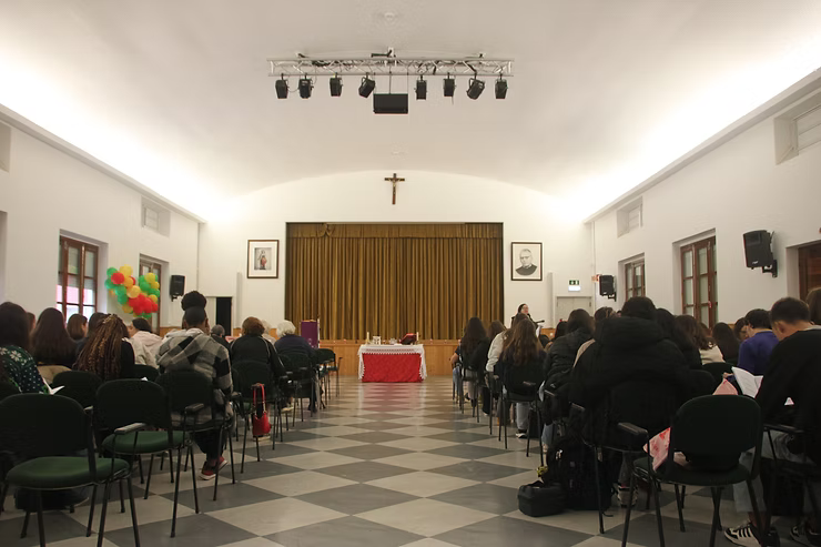 Pessoas sentadas em um pequeno salão de frente para um palco com cortinas, com um pódio vermelho e um crucifixo acima.