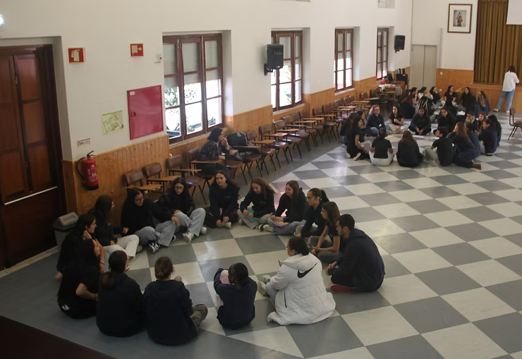 Pessoas sentadas em um grande salão com piso quadriculado, dispostas em círculo e em pequenos grupos próximos às paredes.