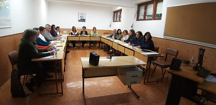 Pessoas sentadas ao redor de mesas em formato de U em uma pequena sala de reuniões, de frente para um palestrante central.