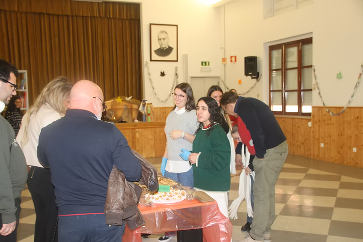 Pessoas reunidas em volta de uma mesa em um salão, conversando e servindo comida em um evento interno.