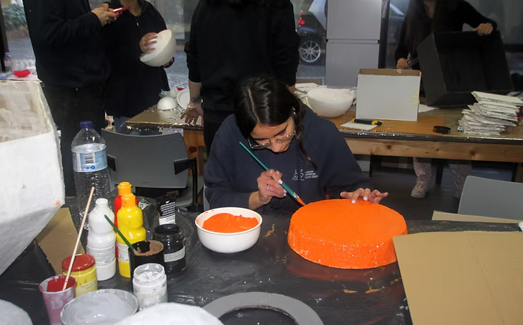 Pessoa pintando uma tigela laranja brilhante em uma mesa com materiais de arte e outras pessoas por perto.