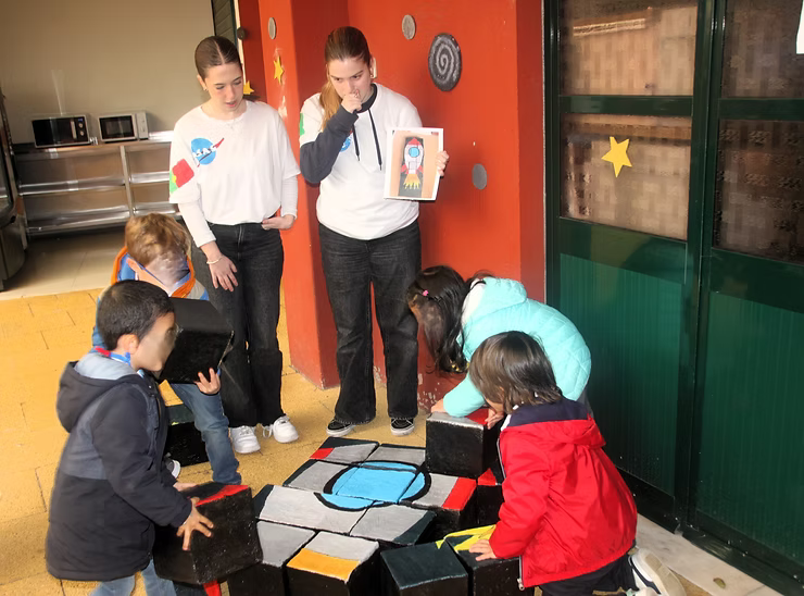 Crianças reunidas em volta de um tapete no chão, com dois adultos por perto, em uma sala de aula ao lado de uma porta verde.