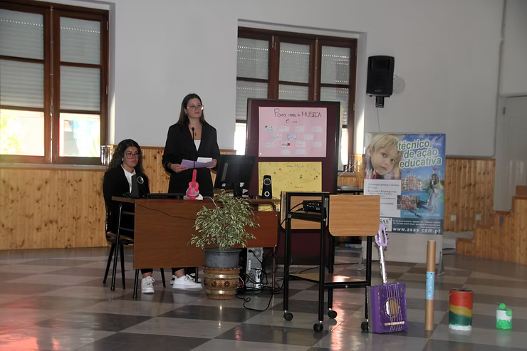 Duas mulheres em um pódio em um salão, uma falando e a outra sentada, com uma tela de apresentação atrás delas.
