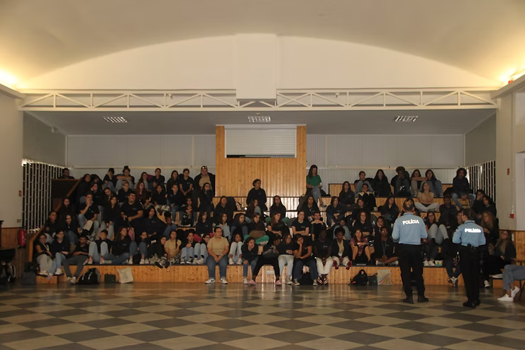 Público sentado em um auditório fechado com dois funcionários uniformizados em pé na frente, sob luzes brilhantes no teto.
